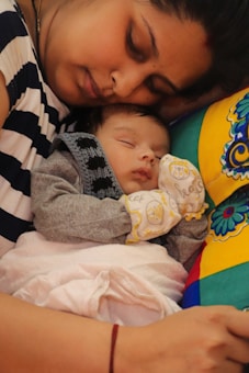 A mother and her baby are peacefully sleeping together. The mother rests her head gently on the colorful pillow, while the baby, dressed in a gray onesie and patterned mittens, is cozy in her arms. The scene is tender and tranquil.