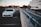 A white car is parked in an empty parking space on the top level of a parking garage. The area appears to be in an urban setting with modern office buildings in the background. There is also a concrete barrier separating the parking area from a street under construction below.
