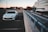 A white car is parked in an empty parking space on the top level of a parking garage. The area appears to be in an urban setting with modern office buildings in the background. There is also a concrete barrier separating the parking area from a street under construction below.