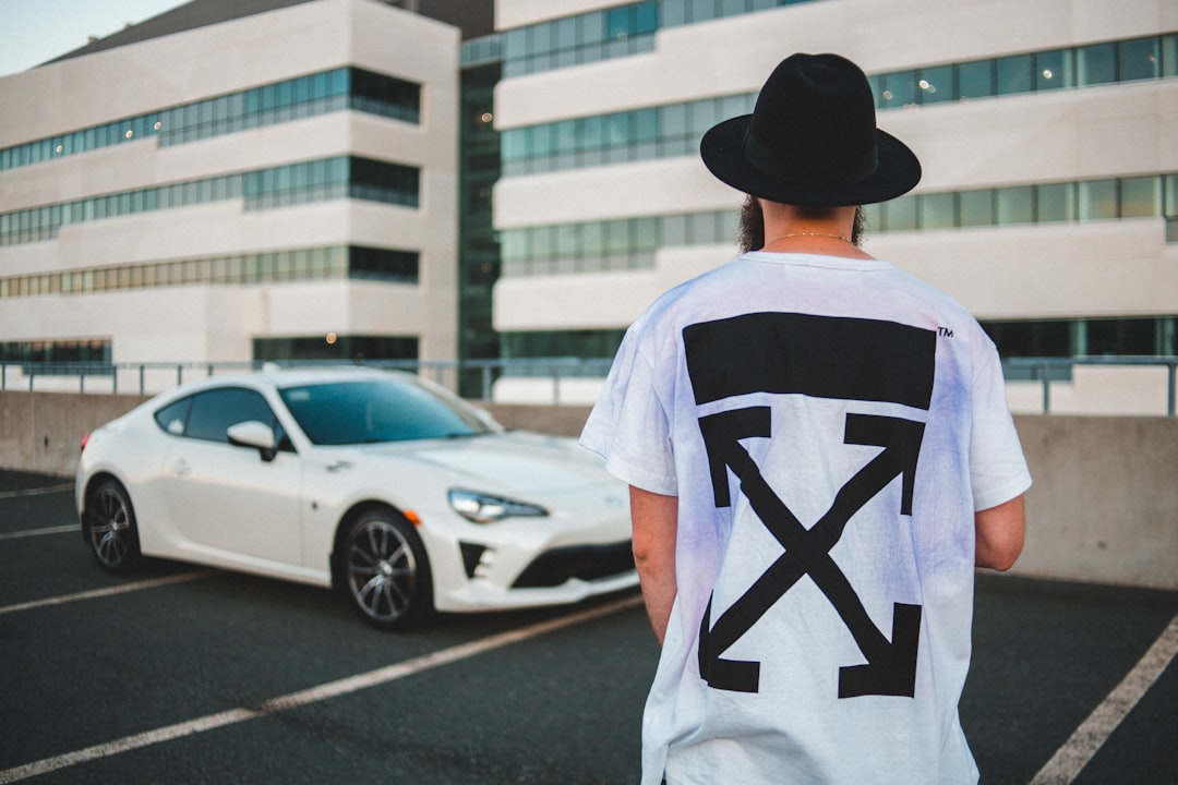 man in white and black t-shirt wearing black fedora hat standing near white sedan during