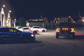 Three cars are parked in a dimly lit area at night, with a blue car on the left, a white car with pink accents in the center, and a dark truck with bright headlights on the right. The vehicles are in an industrial area with a building in the background.