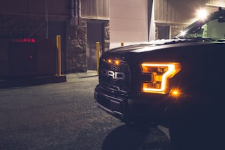 The front of a dark-colored truck is parked in an industrial setting. Bright, amber headlights are illuminated, casting a warm glow. The area is dimly lit, with streetlights contributing faint light and shadows. Industrial buildings and a concrete ground add to the urban feel.