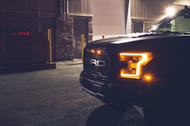 The front of a dark-colored truck is parked in an industrial setting. Bright, amber headlights are illuminated, casting a warm glow. The area is dimly lit, with streetlights contributing faint light and shadows. Industrial buildings and a concrete ground add to the urban feel.