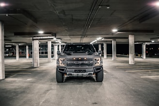 A large black pickup truck is parked in an underground parking garage. The lighting is dim, with overhead lights casting a soft glow on the concrete floor. The truck is centered in the frame, and its headlights are off.