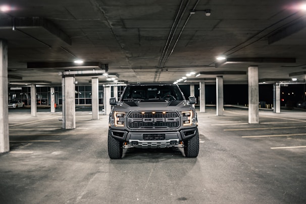 A large black pickup truck is parked in an underground parking garage. The lighting is dim, with overhead lights casting a soft glow on the concrete floor. The truck is centered in the frame, and its headlights are off.