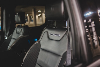 Close-up of a car's interior showcasing a black leather seat with 'Raptor' embroidered on the headrest. The background is slightly blurred, indicating a dimly lit parking area.