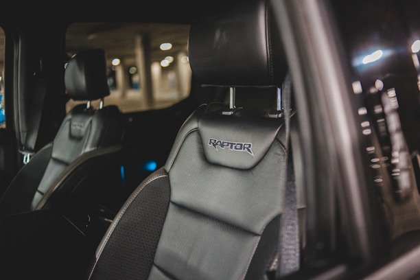 Close-up of a car's interior showcasing a black leather seat with 'Raptor' embroidered on the headrest. The background is slightly blurred, indicating a dimly lit parking area.