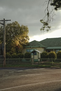 A residential scene featuring a modest house with a green roof and a small front gate surrounded by a low wire fence. The yard is landscaped with hedges and trees, some of which exhibit autumn colors. Overhead power lines and a utility pole are visible against a cloudy sky, with sunlight filtering through.