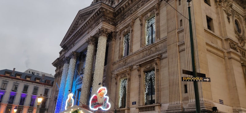 The image features an ornate building adorned with classical architectural elements, including pillars and intricate carvings. Festive lights, resembling icicles, are draped in front of the structure. A neon sign with a Santa Claus and the words 'Hier Gl&uuml;hwein' is prominent in the foreground. The surrounding area shows other buildings with colorful lights, creating a lively atmosphere.