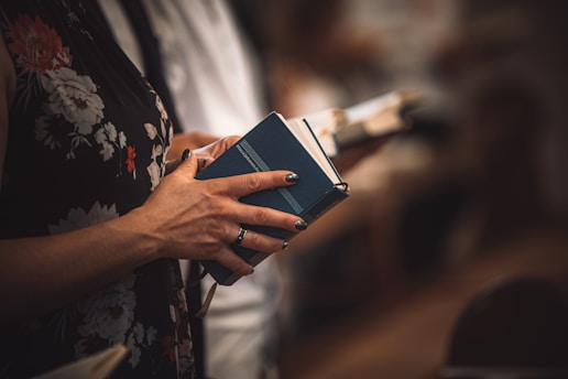 A person holding a book with dark blue cover, possibly in a setting that appears to be formal or religious. The person's fingers have painted nails and rings, and they are wearing a floral-patterned garment. Other people are seen in the background, also holding similar books.