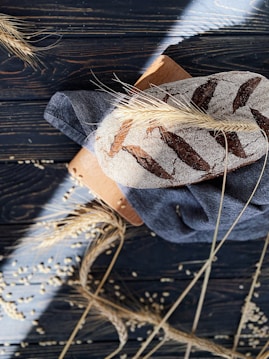 A rustic loaf of bread with a dusting of flour and decorative scoring rests on a dark wooden backdrop. Stalks of wheat and scattered grains surround the bread, adding a natural, artisanal touch. The bread sits atop a blue cloth and a wooden cutting board, with sunlight casting dramatic shadows across the scene.