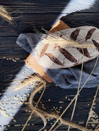 A rustic loaf of bread with a dusting of flour and decorative scoring rests on a dark wooden backdrop. Stalks of wheat and scattered grains surround the bread, adding a natural, artisanal touch. The bread sits atop a blue cloth and a wooden cutting board, with sunlight casting dramatic shadows across the scene.