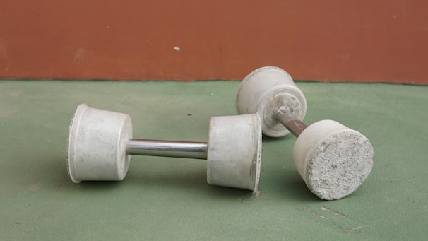 Two white dumbbells with a cylindrical shape lie on a two-toned surface. The surface consists of a green floor and a brown wall in the background. The dumbbells have a metallic handle connecting the weights on either side.