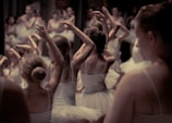 A group of young ballet dancers practice their routines, wearing white tutus and leotards. They are positioned closely together, with their arms raised gracefully in various ballet poses. The background is blurred, focusing attention on the intricate movements of the dancers.
