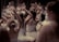A group of young ballet dancers practice their routines, wearing white tutus and leotards. They are positioned closely together, with their arms raised gracefully in various ballet poses. The background is blurred, focusing attention on the intricate movements of the dancers.