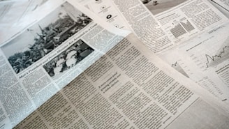 A collection of printed newspapers with articles containing texts, images, and graphs spread out. The layout includes columns of text, black and white photographs, and statistical graphs, conveying information on a flat surface.