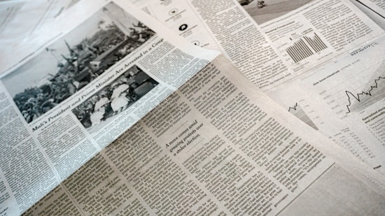 A collection of printed newspapers with articles containing texts, images, and graphs spread out. The layout includes columns of text, black and white photographs, and statistical graphs, conveying information on a flat surface.
