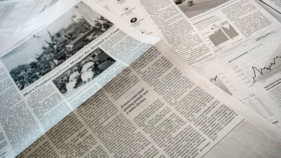 A collection of printed newspapers with articles containing texts, images, and graphs spread out. The layout includes columns of text, black and white photographs, and statistical graphs, conveying information on a flat surface.
