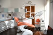 A modern dental office with an orange dental chair in the center, surrounded by various dental equipment such as overhead lights, monitors, and shelves with supplies. The room is well-lit with natural light coming from a window on the right. A clock is mounted on the wall above a wooden door, and an orange chair is placed next to a storage cabinet with a dental instrument sterilizer.