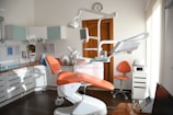 A modern dental office with an orange dental chair in the center, surrounded by various dental equipment such as overhead lights, monitors, and shelves with supplies. The room is well-lit with natural light coming from a window on the right. A clock is mounted on the wall above a wooden door, and an orange chair is placed next to a storage cabinet with a dental instrument sterilizer.