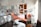 A modern dental office with an orange dental chair in the center, surrounded by various dental equipment such as overhead lights, monitors, and shelves with supplies. The room is well-lit with natural light coming from a window on the right. A clock is mounted on the wall above a wooden door, and an orange chair is placed next to a storage cabinet with a dental instrument sterilizer.