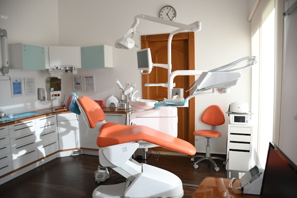 A modern dental office with an orange dental chair in the center, surrounded by various dental equipment such as overhead lights, monitors, and shelves with supplies. The room is well-lit with natural light coming from a window on the right. A clock is mounted on the wall above a wooden door, and an orange chair is placed next to a storage cabinet with a dental instrument sterilizer.