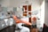 A modern dental office with an orange dental chair in the center, surrounded by various dental equipment such as overhead lights, monitors, and shelves with supplies. The room is well-lit with natural light coming from a window on the right. A clock is mounted on the wall above a wooden door, and an orange chair is placed next to a storage cabinet with a dental instrument sterilizer.