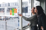 Two women are standing next to a large window, smiling and engaging in discussion. One woman is pointing with a marker at colorful sticky notes displayed on the window. The notes consist of different colors such as orange and green. They appear to be in a modern office setting, with a view of the buildings and balconies outside.