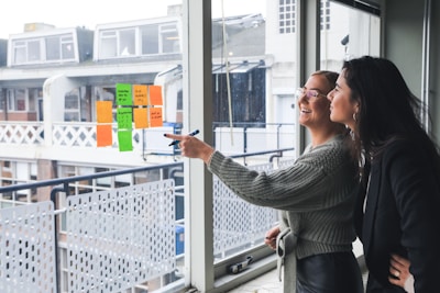 Two women are standing next to a large window, smiling and engaging in discussion. One woman is pointing with a marker at colorful sticky notes displayed on the window. The notes consist of different colors such as orange and green. They appear to be in a modern office setting, with a view of the buildings and balconies outside.