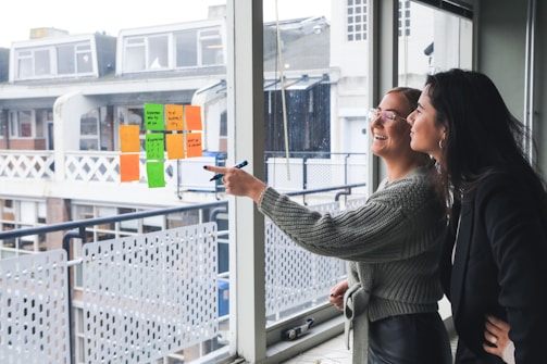 Two women are standing next to a large window, smiling and engaging in discussion. One woman is pointing with a marker at colorful sticky notes displayed on the window. The notes consist of different colors such as orange and green. They appear to be in a modern office setting, with a view of the buildings and balconies outside.