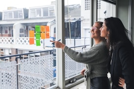 Two women are standing next to a large window, smiling and engaging in discussion. One woman is pointing with a marker at colorful sticky notes displayed on the window. The notes consist of different colors such as orange and green. They appear to be in a modern office setting, with a view of the buildings and balconies outside.