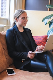A person with glasses is sitting on a brown couch using a laptop, with a red and blue patterned cushion behind them and a mobile phone resting beside them. A potted plant is partially visible in the background, and the setting appears to be indoors with natural light coming through a window.