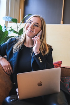 A woman is sitting on a couch with a laptop on her lap, talking on a mobile phone. She is wearing glasses and a black blazer, smiling as she speaks. The setting appears casual with a plant in the background and soft lighting.