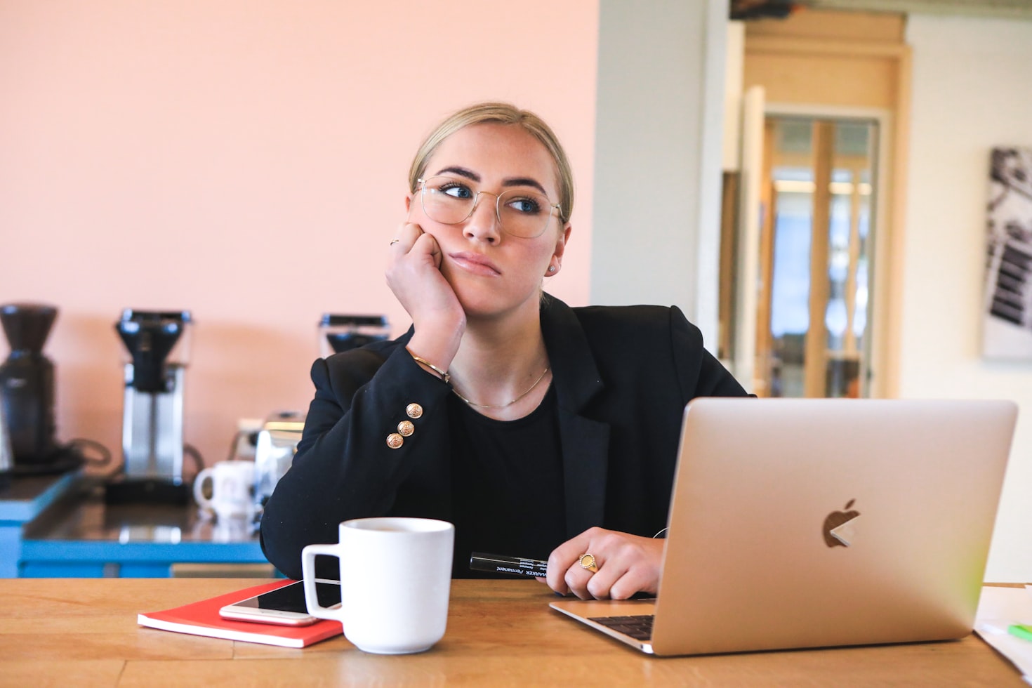 Blonde woman sitting at her desk with a coffee and MacBook, thinking