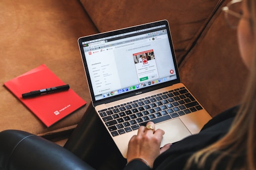 A person is sitting on a couch, using a silver MacBook laptop placed on their lap. The screen displays a webpage with tabs open and various icons visible. Beside the person is a red notebook and a black pen resting on the couch.