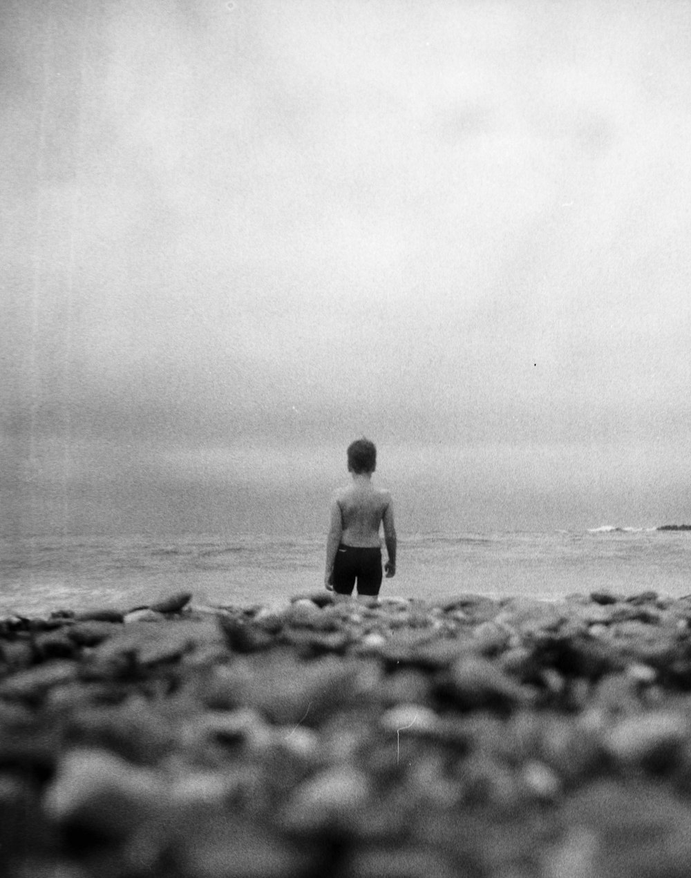 grayscale photo of man standing on rock near body of water