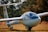 A sleek, white glider resting on a grassy field, with a shiny, glass cockpit and long, slender wings. The reflection of the sky is visible on the cockpit, and a small German flag is painted on the tail fin. The surrounding foliage indicates an autumn setting with vibrant colors.