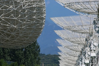 An array of large satellite dishes with intricate metal frameworks is positioned against a backdrop of blue sky and distant mountains. The foreground features lush green trees adding a natural element to the technological scene.