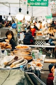 A bustling street market stall features a variety of food dishes, with steam rising from several large pans filled with food. People busy themselves both behind and in front of the stall, engaging in cooking and serving. Brightly lit by hanging bulbs, the scene conveys a sense of lively activity amidst a diverse crowd. Bottles of drink and small plates of dishes are scattered on the counter.