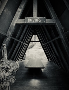 A black and white image of a wooden structure with an A-frame design, featuring a horizontal beam displaying a sign with the word 'BUYUR'. Inside, there is a long wooden table and some plants on the left side. The view through the structure opens up to a serene outdoor scene with a body of water and greenery.
