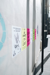 A series of colorful protest posters are affixed to a white wall, each carrying messages like 'We Want to Live' and 'Deeds Not Words'. The posters are aligned horizontally and appear in colors such as yellow, pink, and green. A drawing of a dove with a heart is visible on one of the posters on the left side.