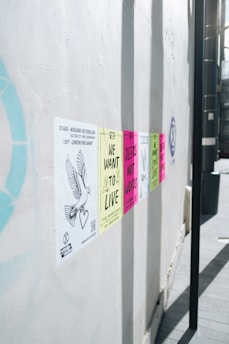 A series of colorful protest posters are affixed to a white wall, each carrying messages like 'We Want to Live' and 'Deeds Not Words'. The posters are aligned horizontally and appear in colors such as yellow, pink, and green. A drawing of a dove with a heart is visible on one of the posters on the left side.