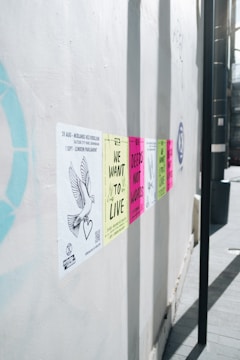 A series of colorful protest posters are affixed to a white wall, each carrying messages like 'We Want to Live' and 'Deeds Not Words'. The posters are aligned horizontally and appear in colors such as yellow, pink, and green. A drawing of a dove with a heart is visible on one of the posters on the left side.