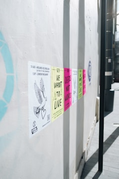 A series of colorful protest posters are affixed to a white wall, each carrying messages like 'We Want to Live' and 'Deeds Not Words'. The posters are aligned horizontally and appear in colors such as yellow, pink, and green. A drawing of a dove with a heart is visible on one of the posters on the left side.