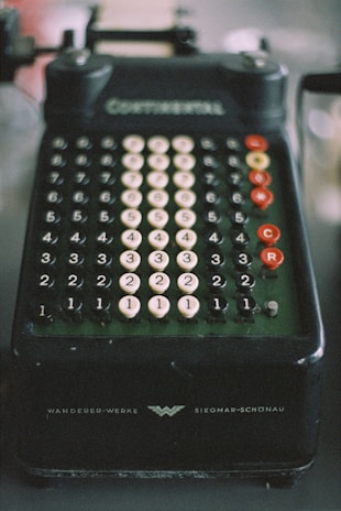 A vintage mechanical calculator with multiple rows of numbered, circular keys. The device is dark in color with white and red keys, and features inscriptions indicating brand details. The overall appearance suggests an era of pre-digital technology.