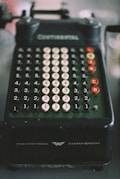 A vintage mechanical calculator with multiple rows of numbered, circular keys. The device is dark in color with white and red keys, and features inscriptions indicating brand details. The overall appearance suggests an era of pre-digital technology.