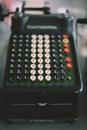 A vintage mechanical calculator with multiple rows of numbered, circular keys. The device is dark in color with white and red keys, and features inscriptions indicating brand details. The overall appearance suggests an era of pre-digital technology.