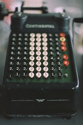 A vintage mechanical calculator with multiple rows of numbered, circular keys. The device is dark in color with white and red keys, and features inscriptions indicating brand details. The overall appearance suggests an era of pre-digital technology.