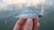 A hand is holding a fishing lure designed to resemble a small fish. The lure has a metallic hook on its back and features a realistic fish design with a clear plastic lip at the front. The background is slightly blurred, showing a body of water in subdued colors.