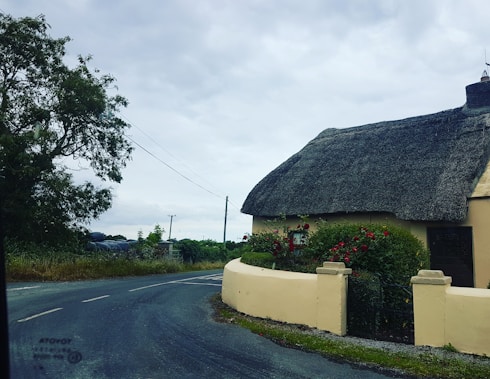 A traditional thatched-roof cottage situated by a rural roadside. The cottage is surrounded by a lush garden with vibrant red flowers and greenery. A curved stone wall separates the garden from the road. The sky is cloudy, and there is a tree and bushes visible in the background.